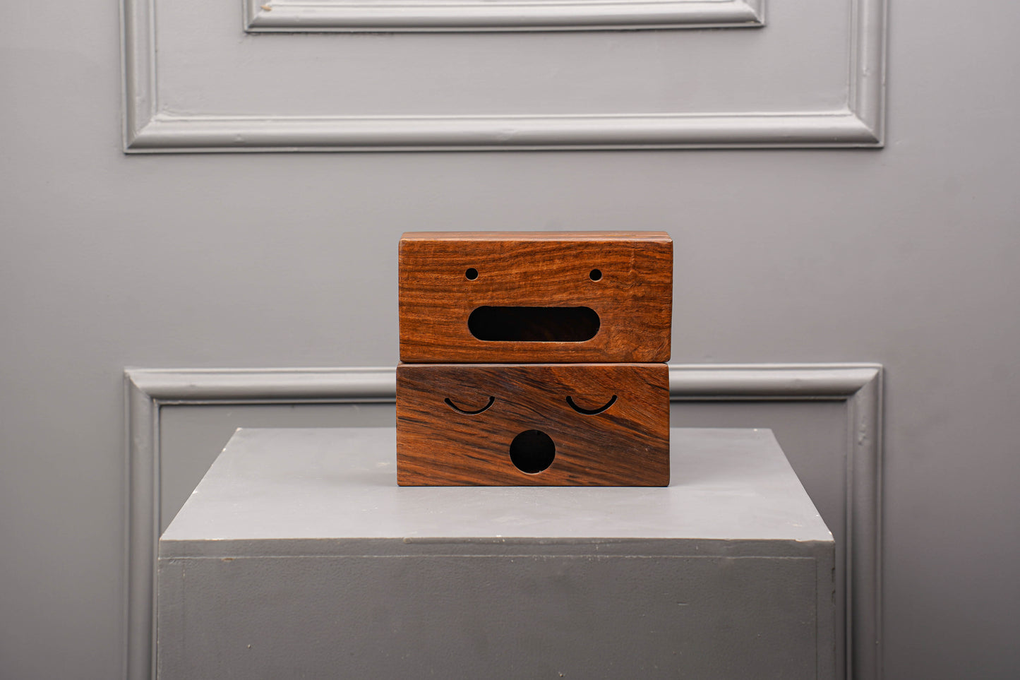 Wooden box with facial features on a gray surface against a gray wall.