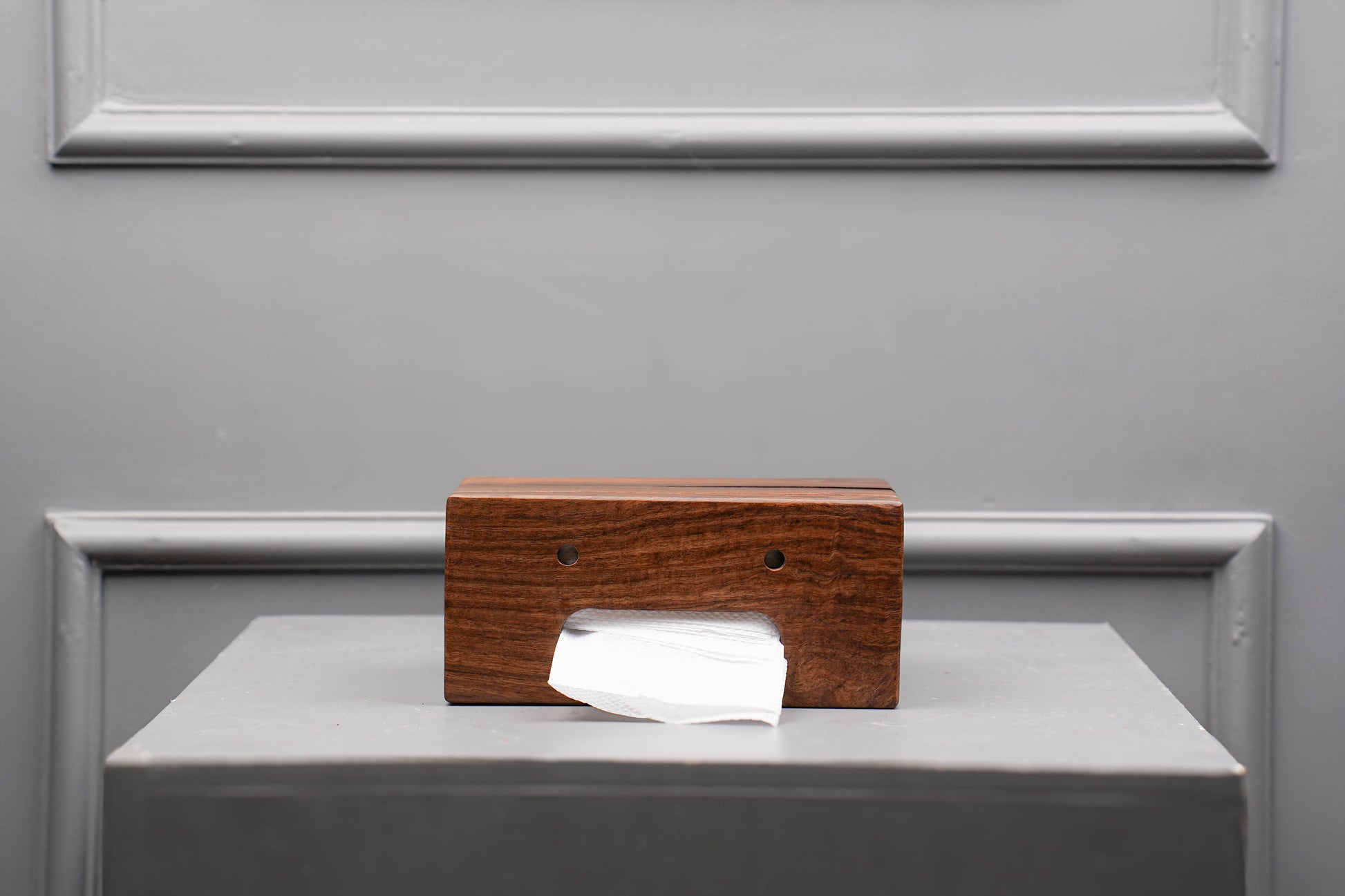 Wooden tissue box with a white tissue on a gray surface against a gray wall.